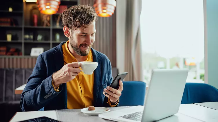 Young Caucasian bearded businessman dressed smart casual using smart phone and drinking coffee while sitting in cafe.