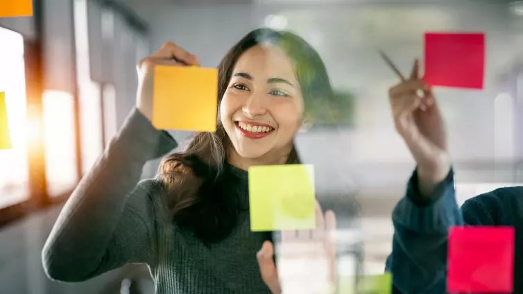 Smiling woman, writing on a post-it note, that's stuck on a glass wall.