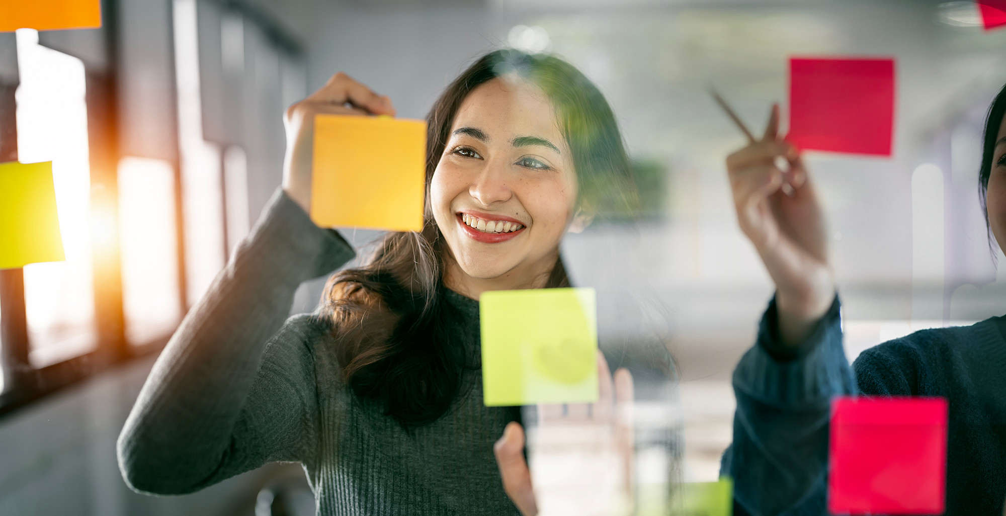 Smiling woman, writing on a post-it note, that's stuck on a glass wall.